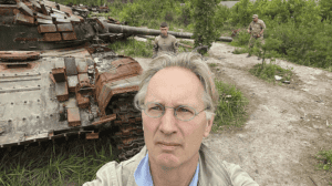 Jaap Scholten selfie in front of a destroyed tank with military personnel in the background, during the Ukraine war.