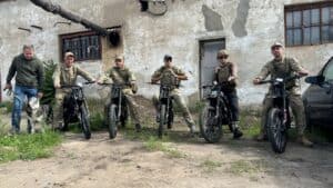 Four Ukrainian soldiers on donated Protect Ukraine electric motorcycles, with a man and a dog standing alongside them.